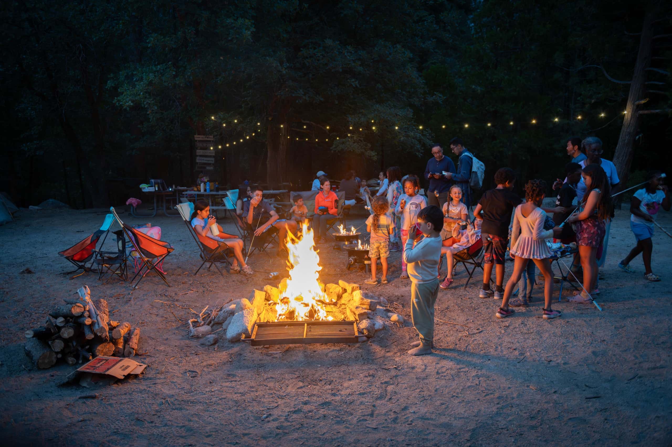 Outdoorithm families gathered around a tall campfire at dusk under string lights, Kings Canyon