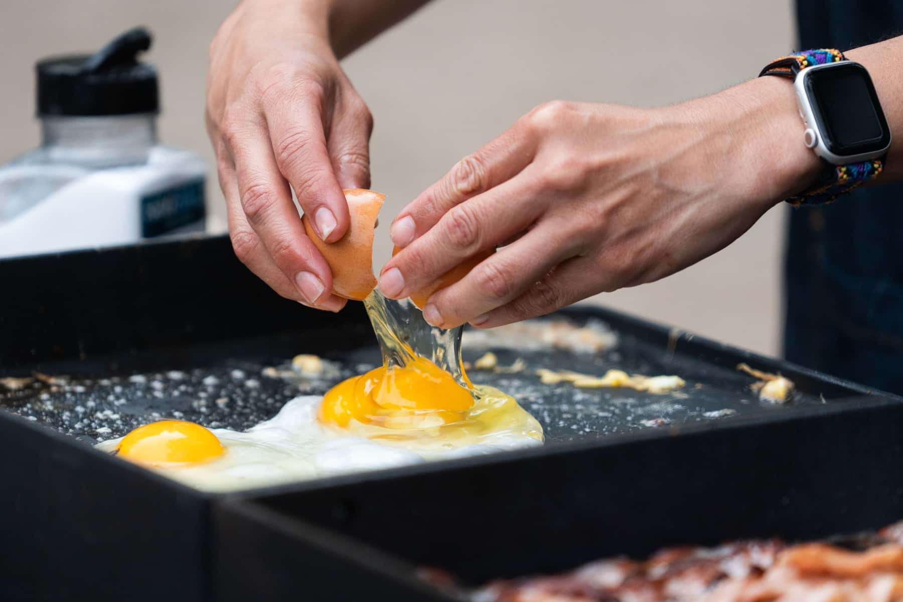 Hands cracking eggs onto the camp griddle beside strips of bacon