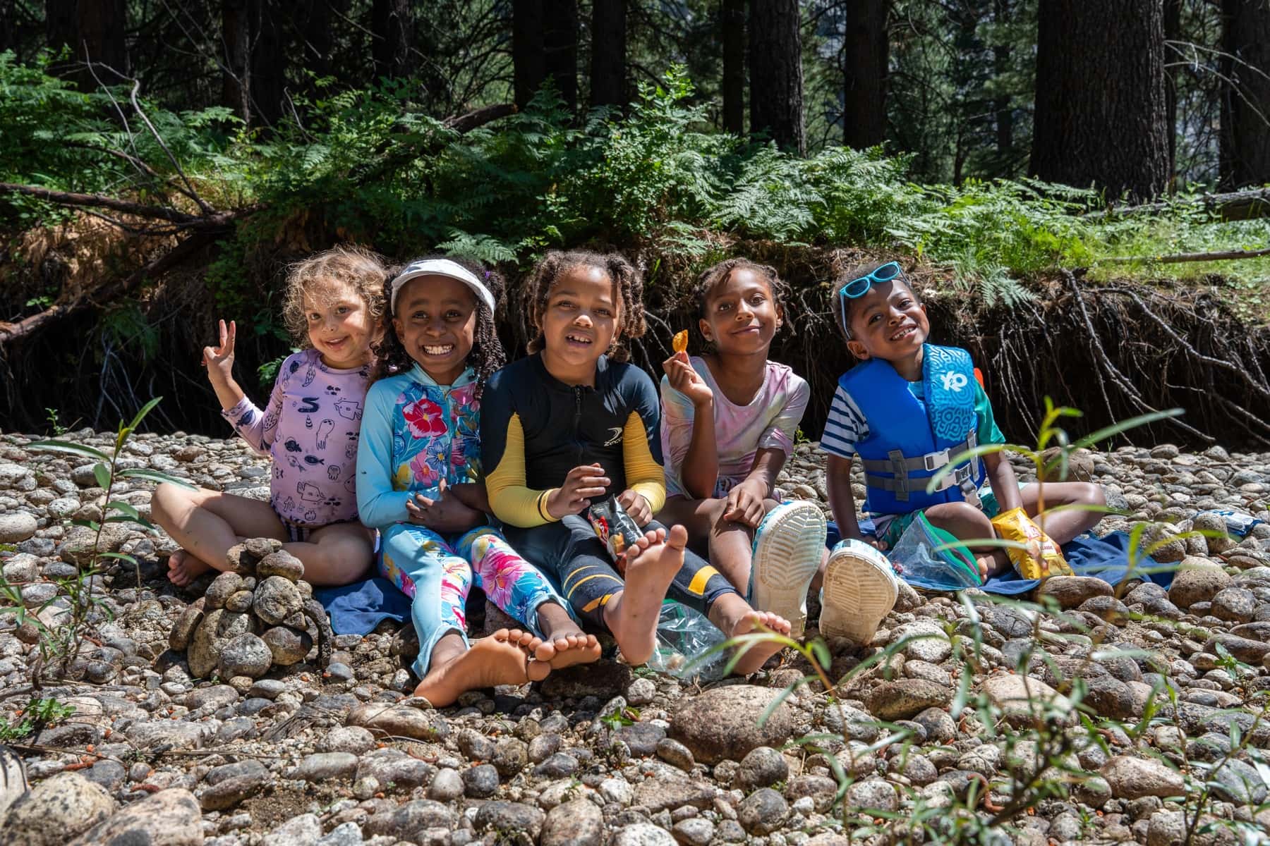 Five children sitting shoulder to shoulder on the riverbank cobbles, all grinning at the camera