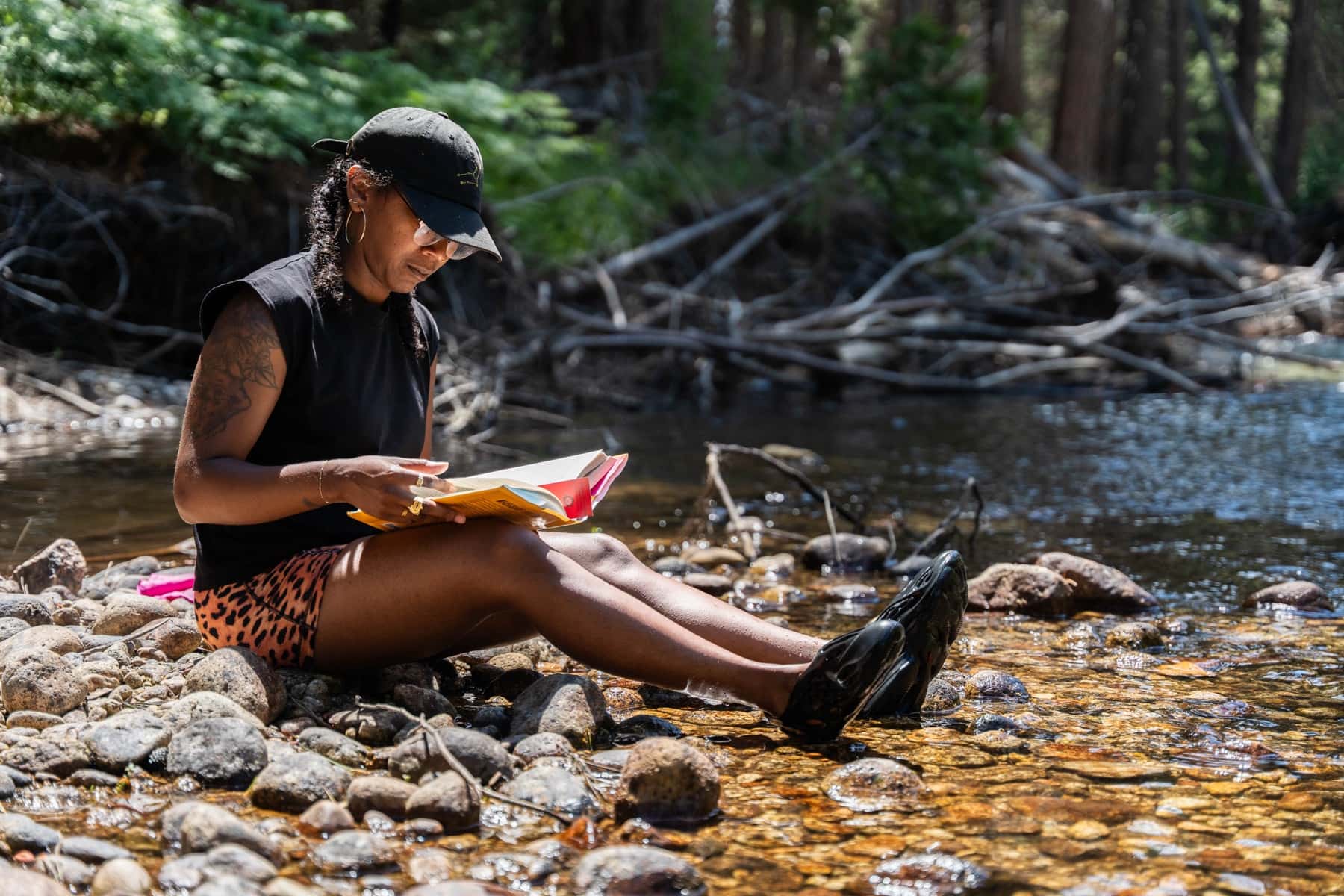 A woman reads a book on the rocky bank with her feet in the stream