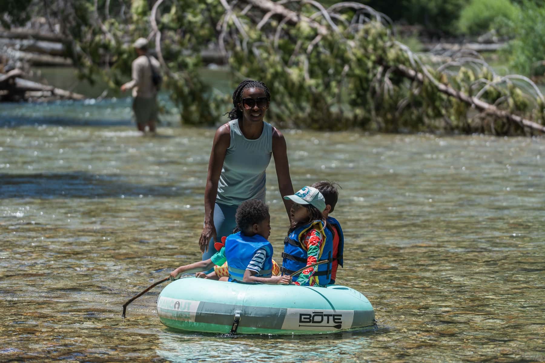 A mother wades a shallow river pushing an inner tube carrying two small children in life vests