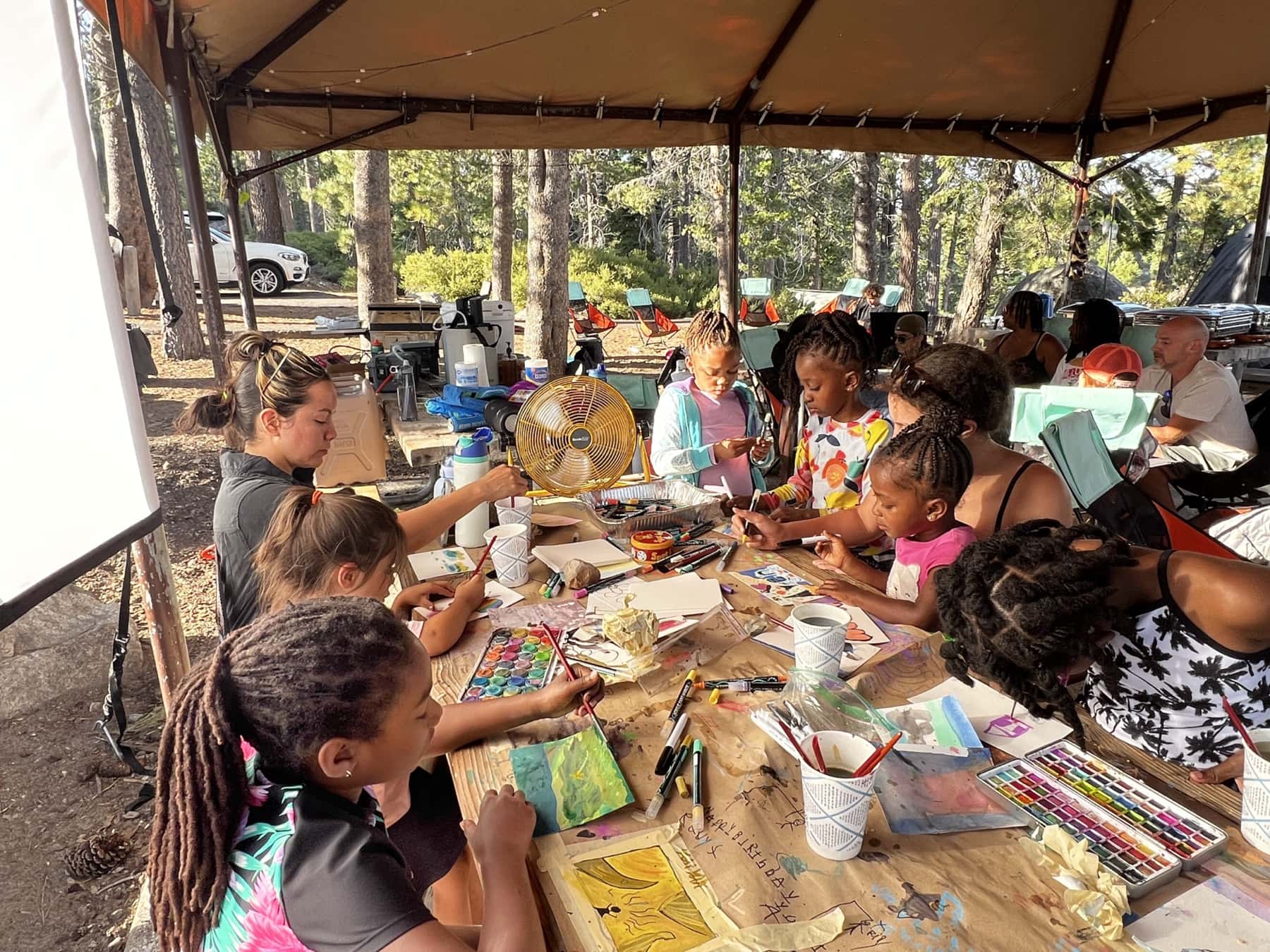 Kids and adults painting with watercolors at a long picnic table under the camp canopy