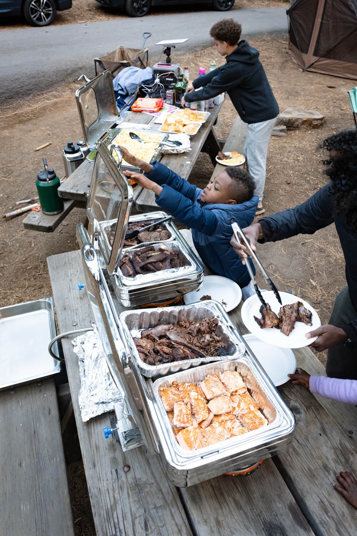 A boy reaches across the camp kitchen table to scoop mac and cheese while an adult serves brisket onto a plate, with chafing trays of brisket and salmon lined up along the picnic table