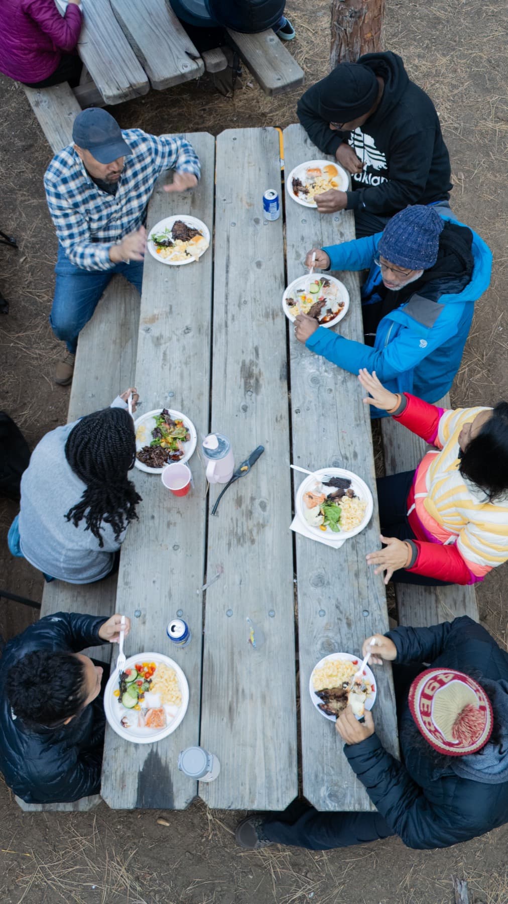 Overhead view of eight adults and children eating plates of brisket and mac and cheese together at a long camp picnic table
