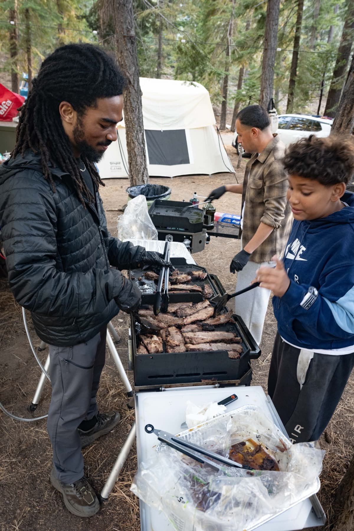Two adults and a teenager searing slices of pre-smoked brisket on the camp flat-top, a cooler of vacuum-sealed bags open in front of them and a canvas wall tent behind
