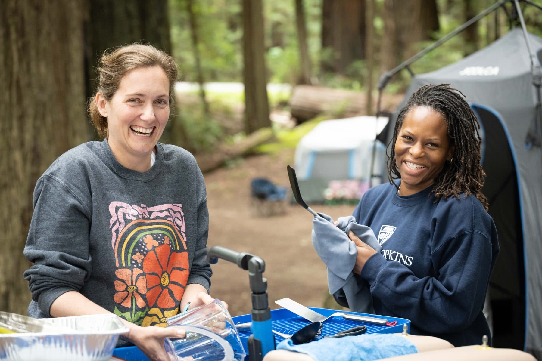 Two campers laughing together at the wash station, drying dishes over the blue wash bins after a meal