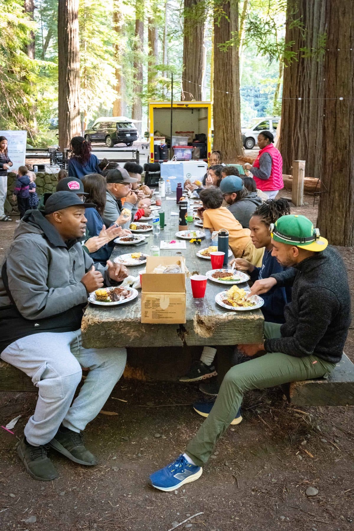 Adults and children eating dinner together at a long picnic table among the redwoods