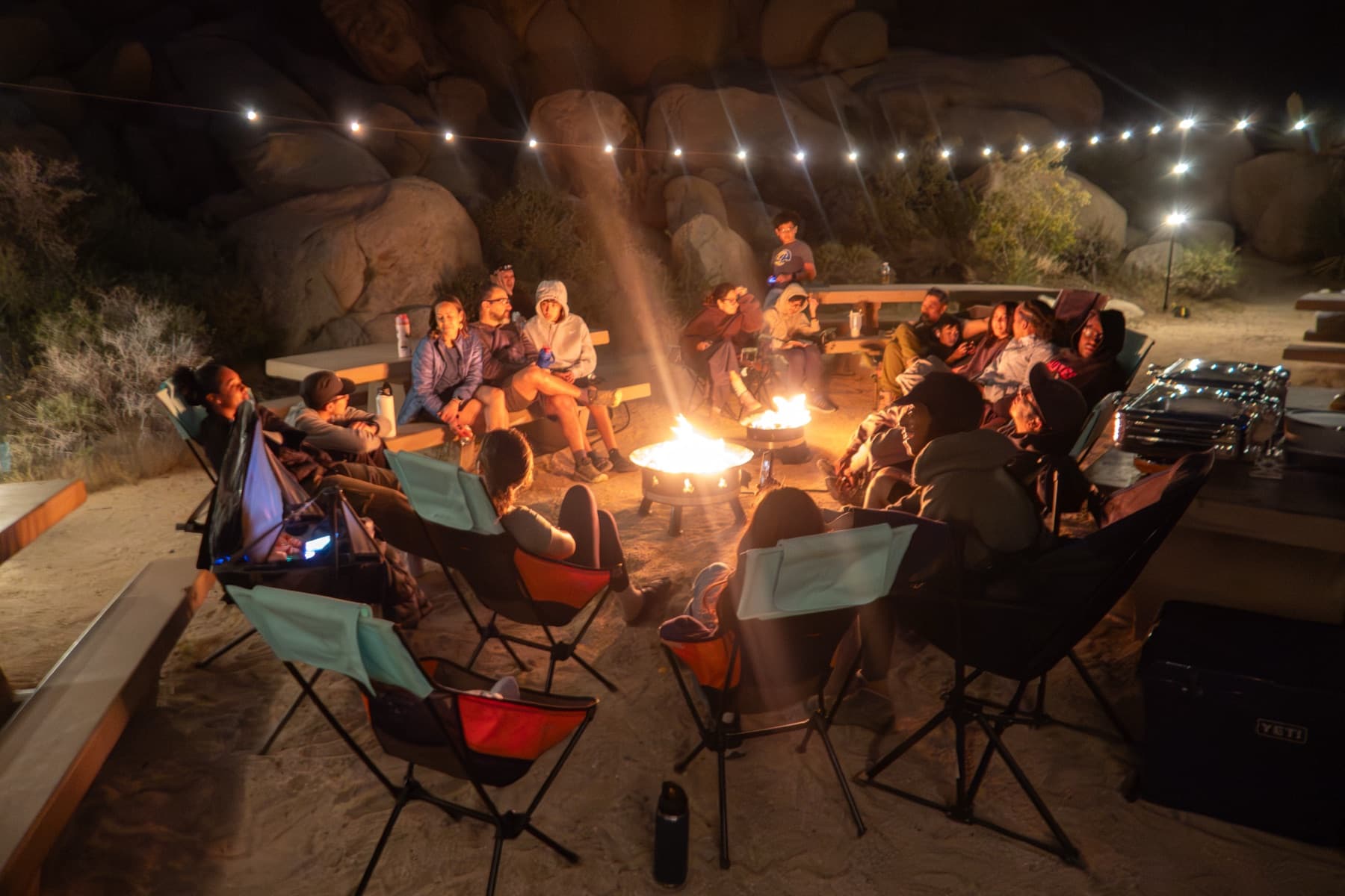 Families gathered in camp chairs around the fire under string lights at Joshua Tree
