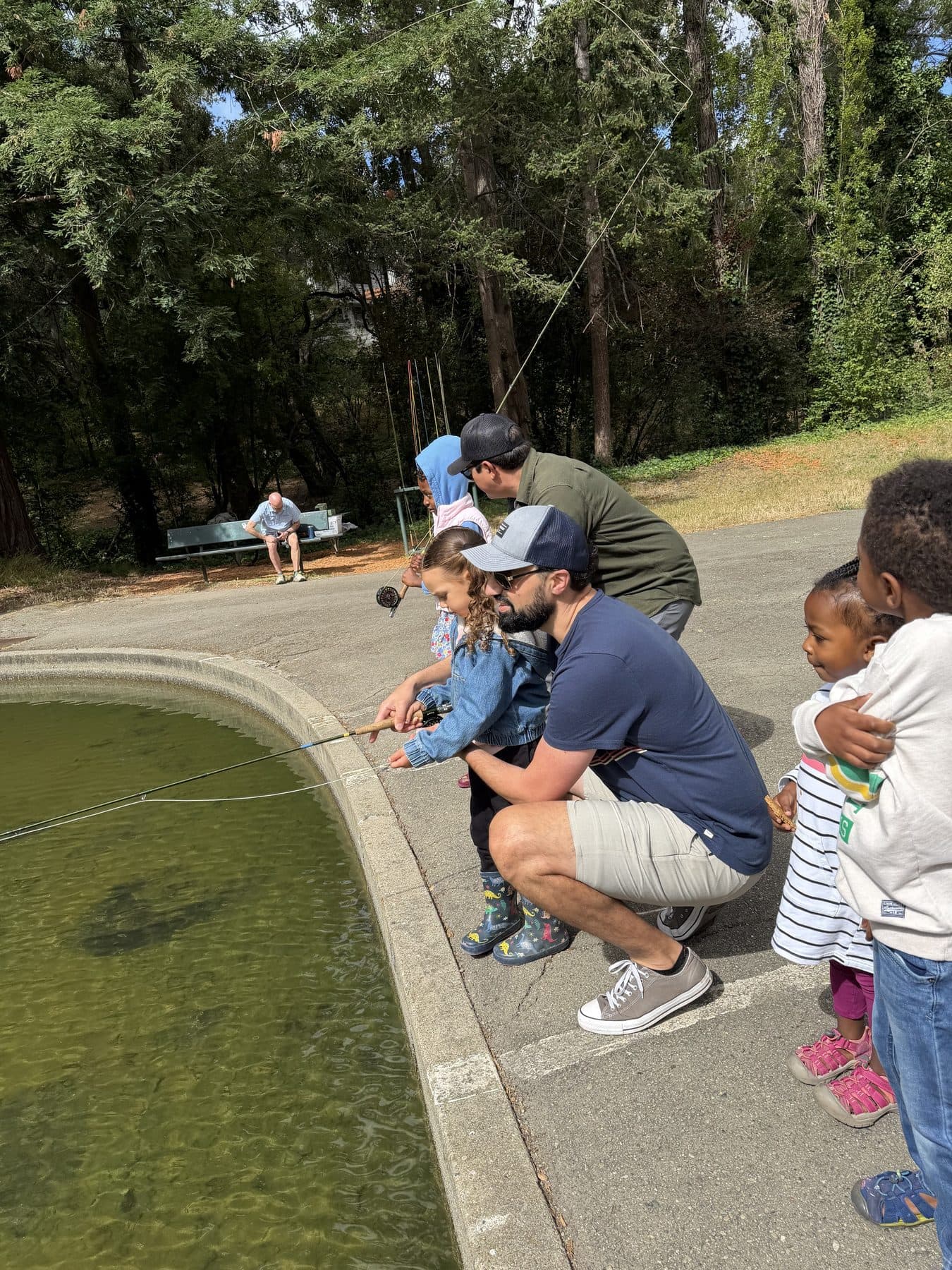 An adult crouches at the pond edge teaching a small child to cast a fly rod while other kids watch
