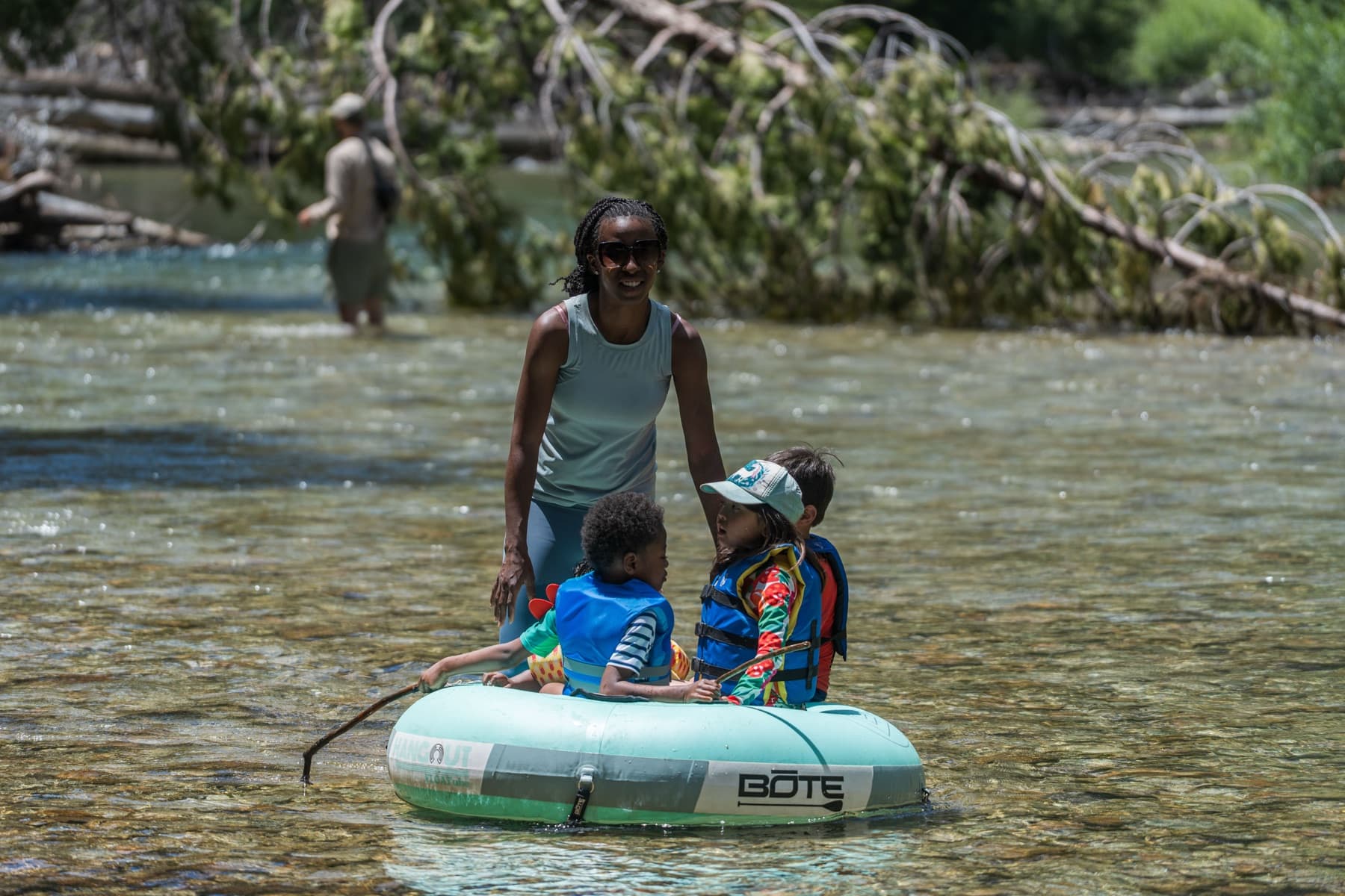 A mother wades a shallow river pushing an inner tube carrying two small children in life vests