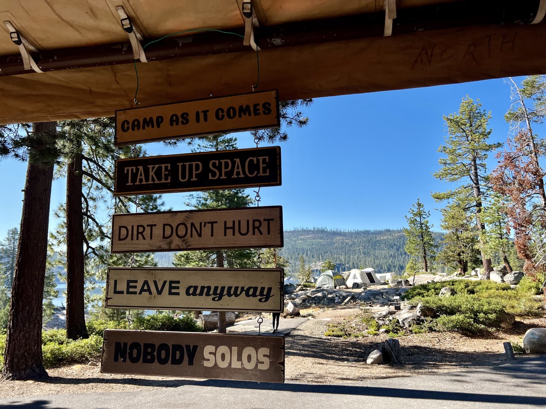 Five hand-painted wooden signs hanging at camp reading Camp As It Comes, Take Up Space, Dirt Don't Hurt, Leave Anyway, and Nobody Solos