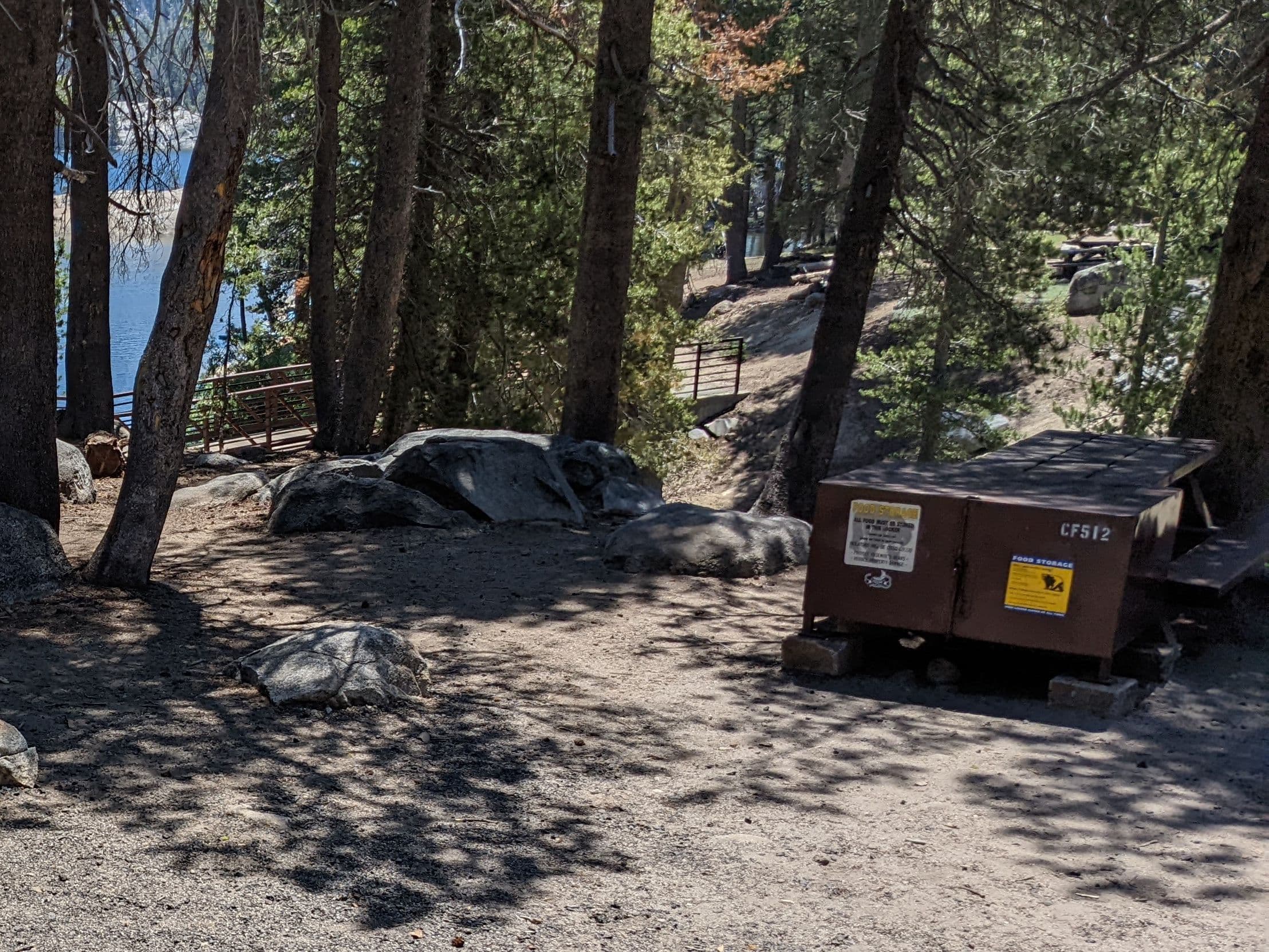 A brown steel bear locker at the edge of a lakeside campsite among pines and granite boulders