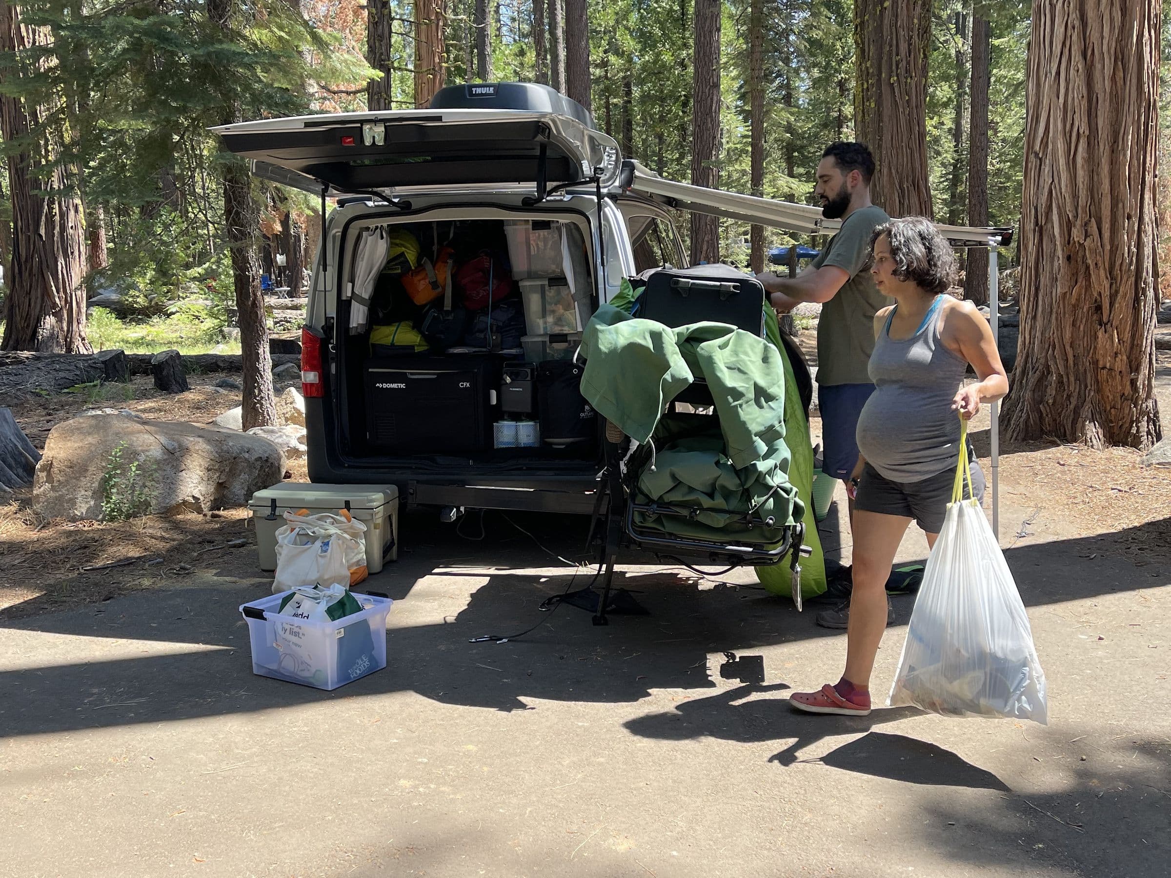 Two campers loading up the open back of their van at a campsite — a cooler and latching bins on the ground, duffel bags stacked inside
