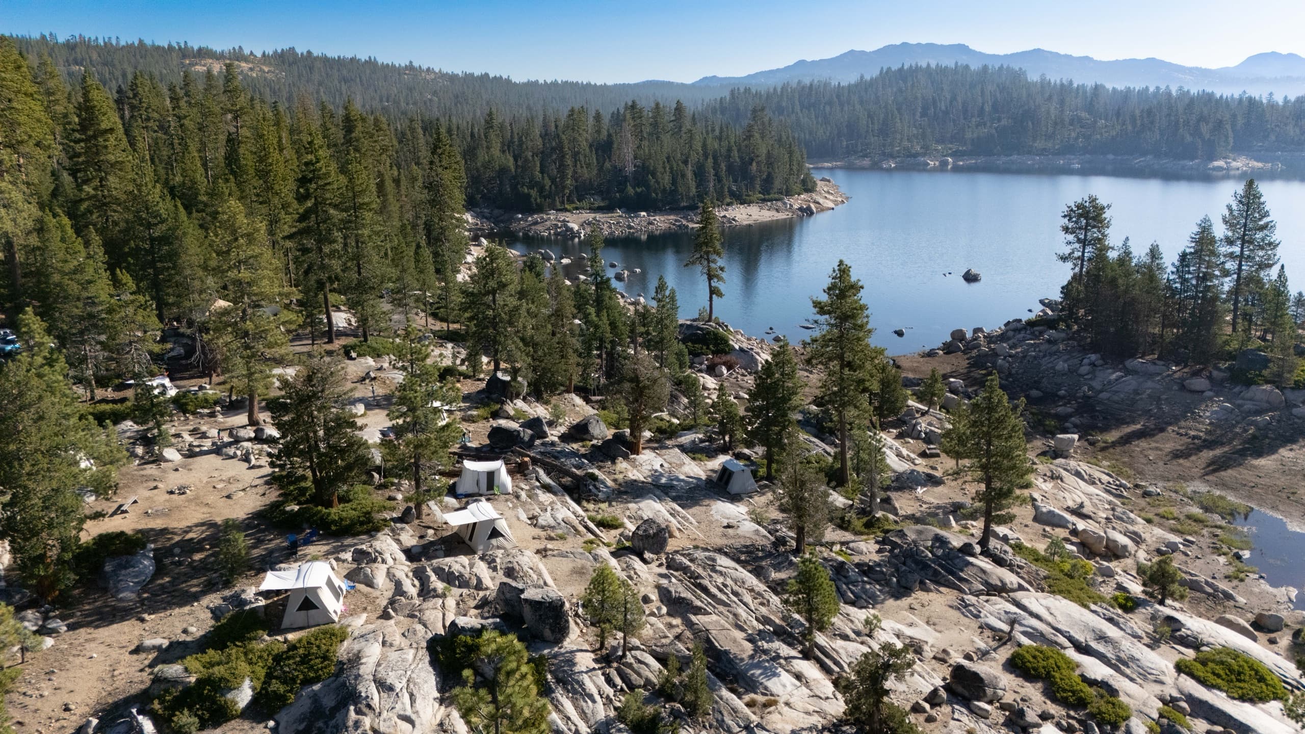 Aerial view of the Outdoorithm Collective tent village pitched on granite slabs above Spicer Reservoir, with forested ridges beyond